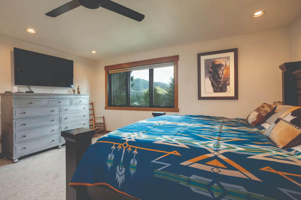 This image presents a tranquil bedroom featuring a large bed dressed with a patterned blanket, a sizable window revealing a stunning mountain view, a flat-screen TV, and bison-themed artwork. The dresser provides ample storage, and the overall atmosphere is serene and comfortable.