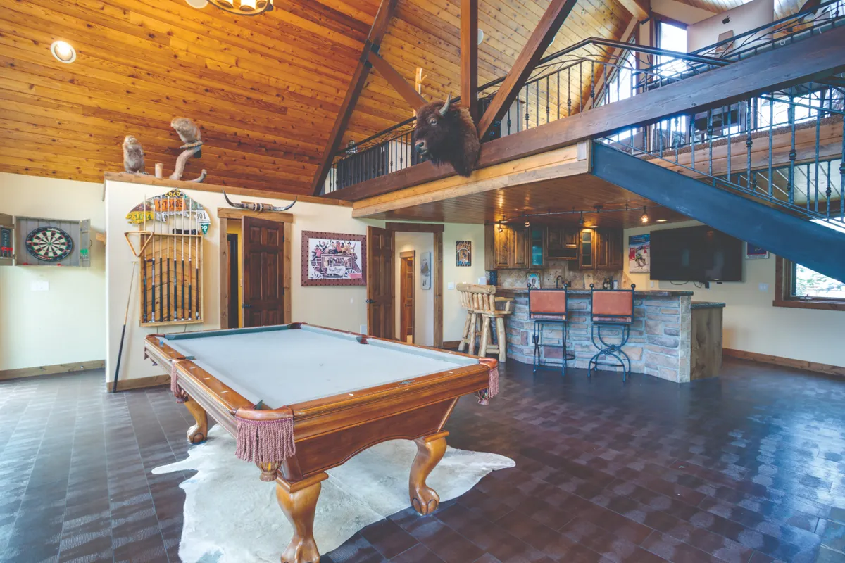 A cozy, rustic game area with a pool table, classic bar seating, and unique decor including a buffalo mount and dartboard. Exposed wooden beams add warmth, and the layout encourages memorable game nights and casual celebrations.