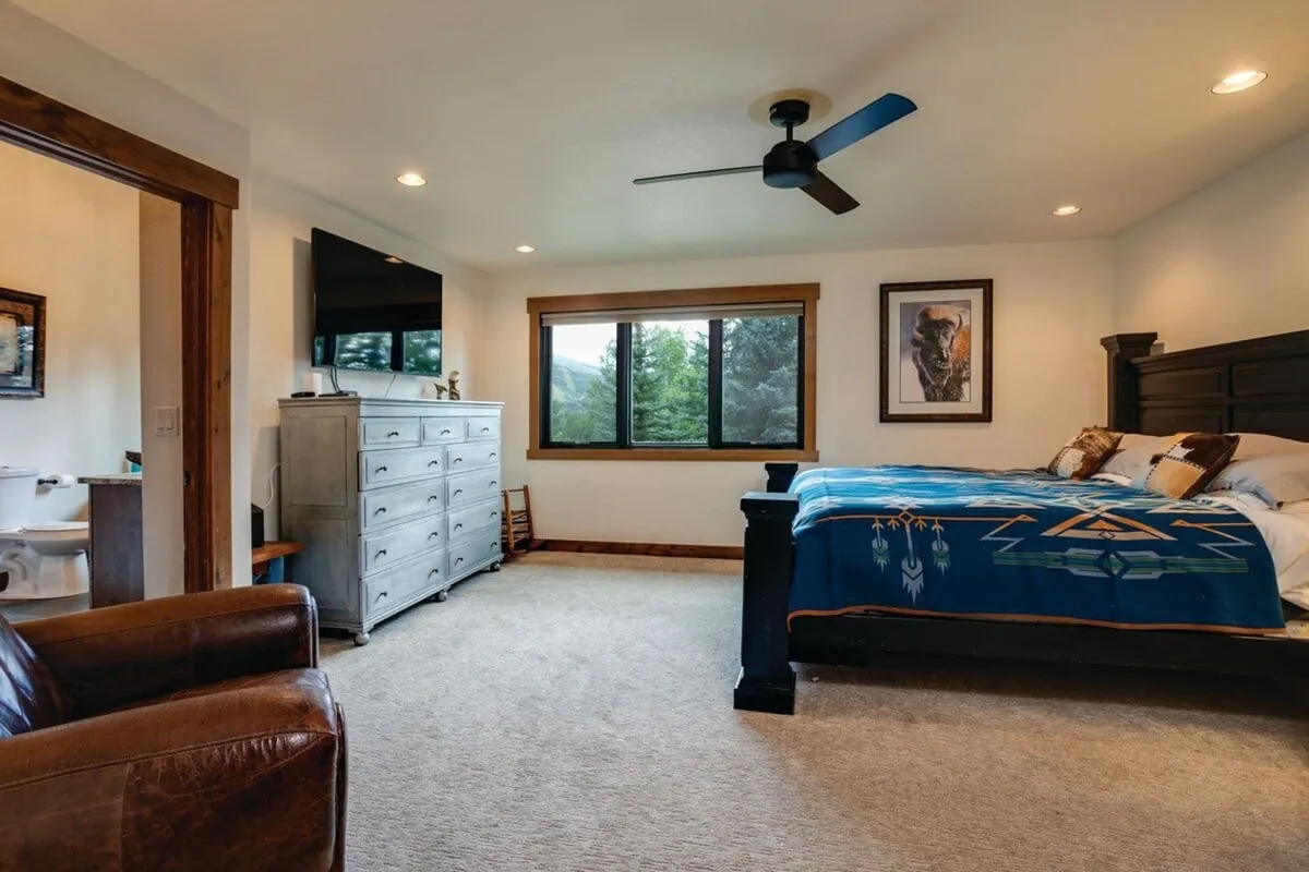 A calm, serene bedroom is depicted here, with a large, plush bed adorned with a decorative blanket, a window offering a view of treetops, a dresser with multiple drawers for storage, and a leather chair for relaxing or reading. The atmosphere is peaceful and inviting, ideal for downtime.