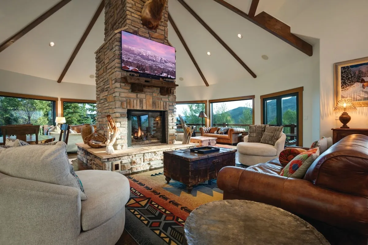 The image showcases a spacious great room with a towering stone fireplace at its center, surrounded by plush leather seating and a vibrant area rug. Expansive windows frame breathtaking mountain views, filling the room with natural light and offering a glimpse of Steamboat Springs' stunning landscapes. The inviting space is perfect for cozy gatherings, movie nights, and memorable family moments.