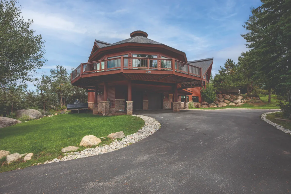 A unique octagonal home with a wrap-around deck, natural stone accents, and a lush green lawn. The architecture stands out while the expansive deck suggests space for hosting large parties or simply relaxing as a group.