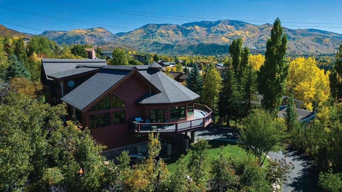 An exterior shot showcasing the house’s distinctive architecture nestled among mature trees, with a dramatic mountain backdrop. The spacious deck is clearly visible, inviting guests to relax and take in the surrounding scenery.