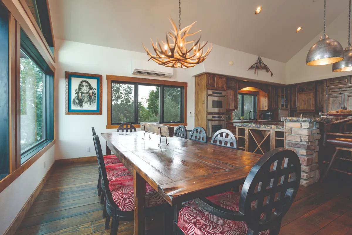 A large wooden dining table sits beneath an elegant antler chandelier, positioned next to massive windows that flood the room with natural light. Framed artwork and rustic decor create a welcoming, celebratory dining atmosphere ideal for gatherings.