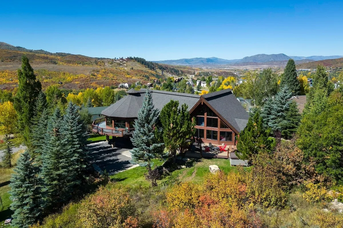 This image features the Moose Meadows Lodge set amid surrounding trees with distant mountains in the background. The home's wooden architecture blends seamlessly with the natural environment, and the deck provides clear views for enjoying sunsets or beginning an outdoor adventure. The tranquil setting hints at the proximity to hiking trails and mountain experiences.