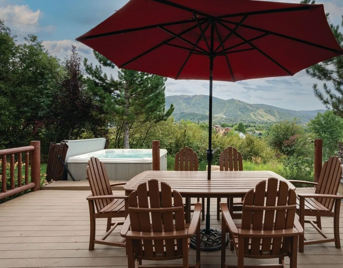 An inviting outdoor space showcasing a wooden dining table under a red umbrella, a hot tub, and breathtaking mountain views. The area is perfectly arranged for al fresco dining, soaking, and lively socialization with friends and family.