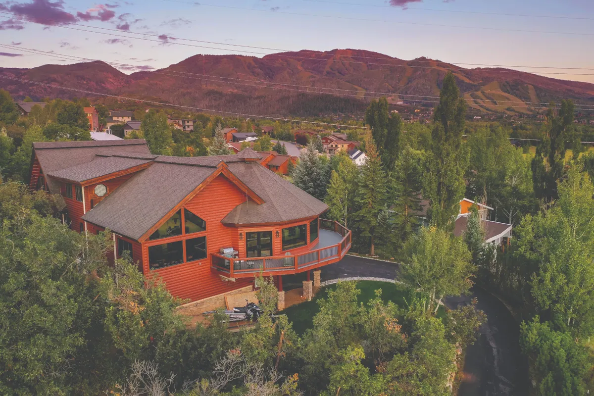This image showcases the lodge's exterior, highlighting its spacious deck, large windows, and the surrounding mountain landscape. The scene is tranquil, with sunlight accentuating both the house and natural surroundings, emphasizing the connection between luxury accommodations and Steamboat's natural beauty.
