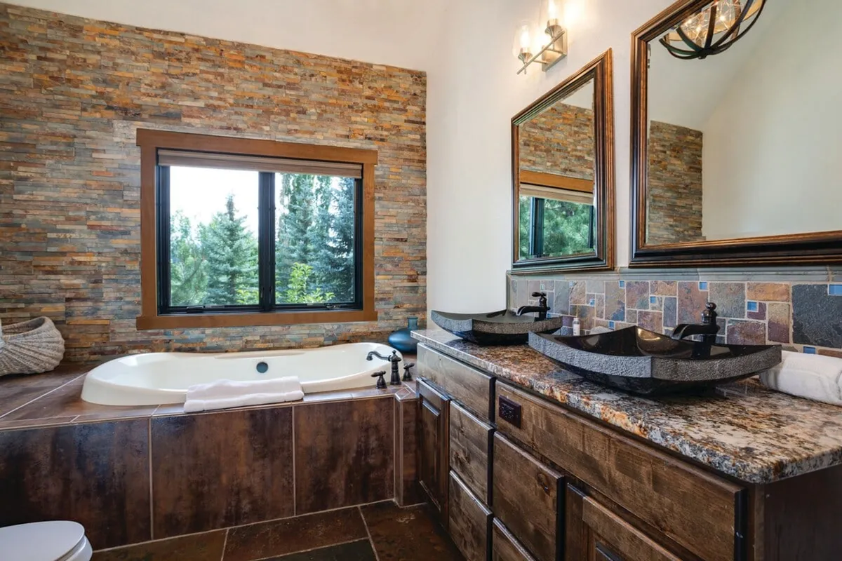 This bathroom exudes spa-like luxury, featuring a large soaking tub beside a stone wall, dual sinks, and a sizable window that lets in natural light. The room balances practicality and indulgence, perfect for unwinding at the end of the day.