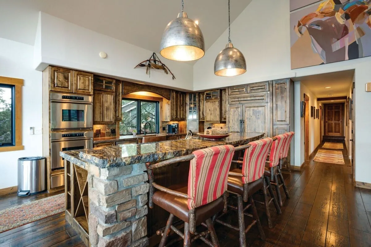 A warmly lit kitchen centered around a substantial granite countertop, with custom wooden cabinetry, striped bar stools, and modern pendant lights. The kitchen exudes an inviting atmosphere ideal for both cooking and entertaining.