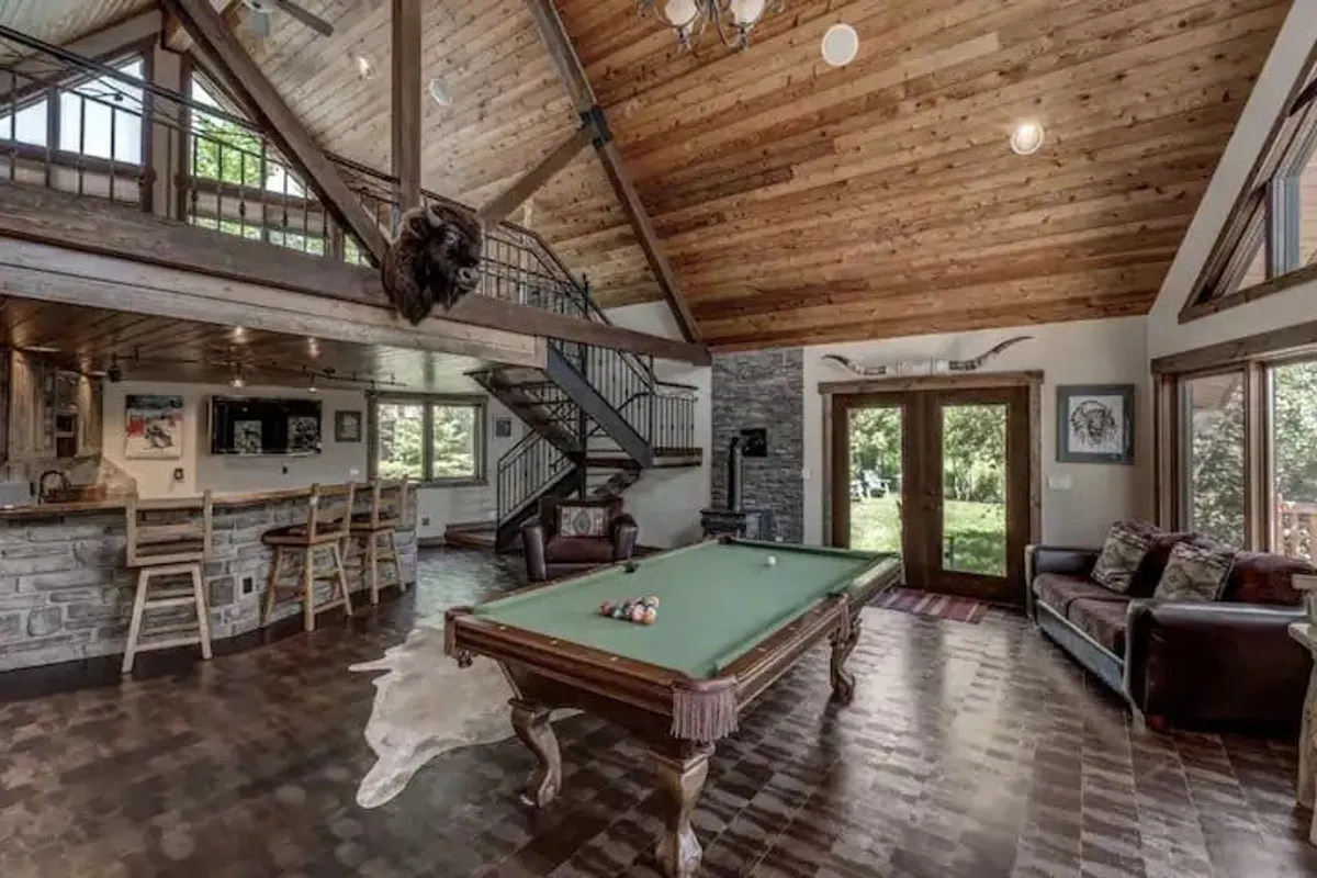 A vibrant game room featuring a pool table, bar area, and large windows flooding the space with light. There's ample seating for guests, and the bar setup encourages interactive and festive gatherings. The overall mood is lively and relaxed, perfect for games, socializing, and enjoying drinks with friends or family.