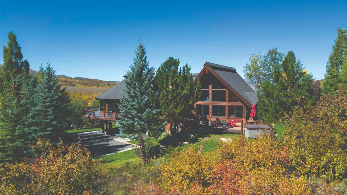 A striking exterior shot of a large wooden lodge nestled amid trees, featuring an expansive deck area with comfortable outdoor furniture. The scene exudes tranquility and hints at ample space for gatherings, barbecues, and relaxation against a natural backdrop.