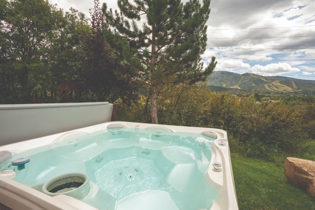 A tranquil hot tub sits on the edge of the property, offering sweeping mountain views and surrounding greenery. The inviting setting suggests a perfect place to relax after skiing, hiking, or cycling, with sunset lighting highlighting the outdoor experience.