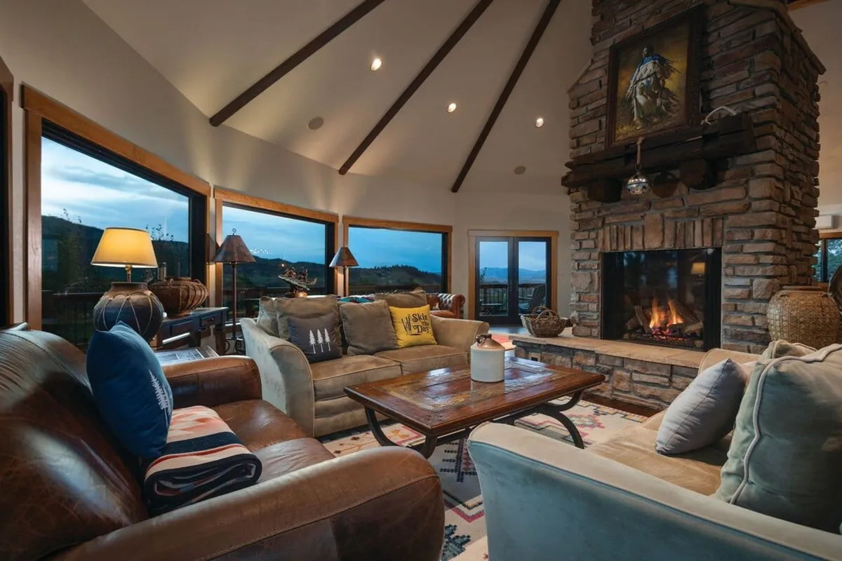 A spacious great room with a welcoming fireplace as the focal point, surrounded by comfortable sofas, a large wooden coffee table, and expansive windows opening up to breathtaking alpine views. The natural light fills the space, blending the cozy warmth of the fire with the grandeur of the surrounding scenery.