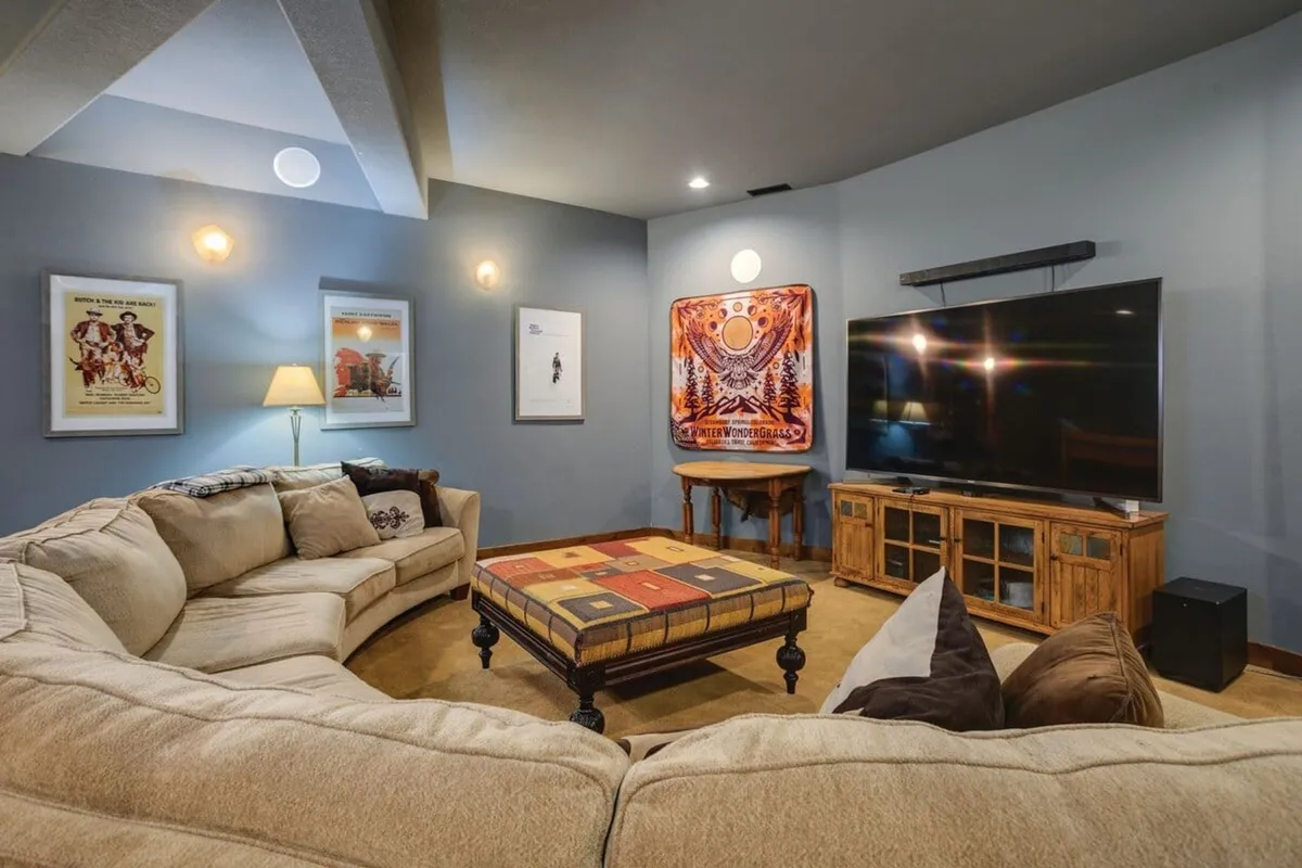 This interior scene features a large sectional sofa arranged for socializing and movie viewing. A colorful ottoman sits nearby, and a TV is visible across the room, with several pieces of vibrant artwork adorning the walls. The layout encourages group gatherings and relaxed entertainment, making it suitable for both movie nights and game sessions.