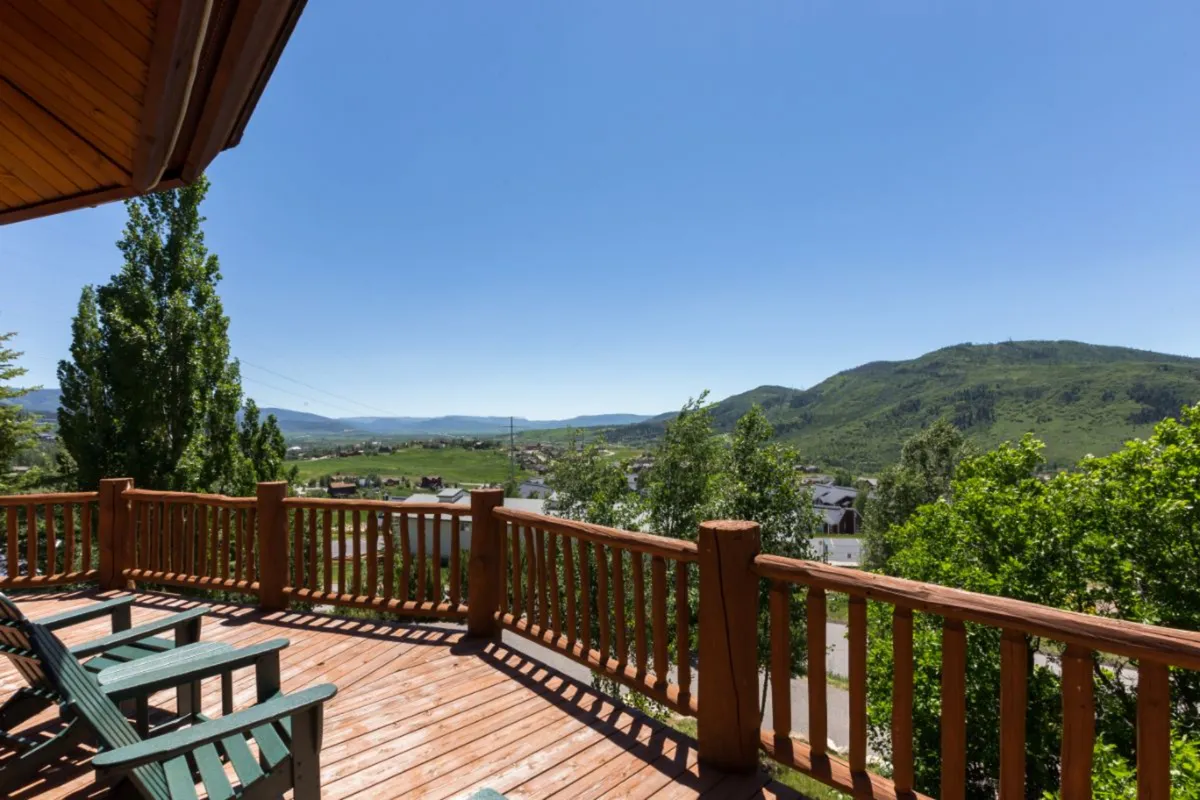 An inviting wooden deck overlooking lush greenery and majestic mountain views, with comfortable outdoor seating that encourages relaxation and socializing. The blue sky and scenic backdrop add to the sense of tranquility and connection to nature.