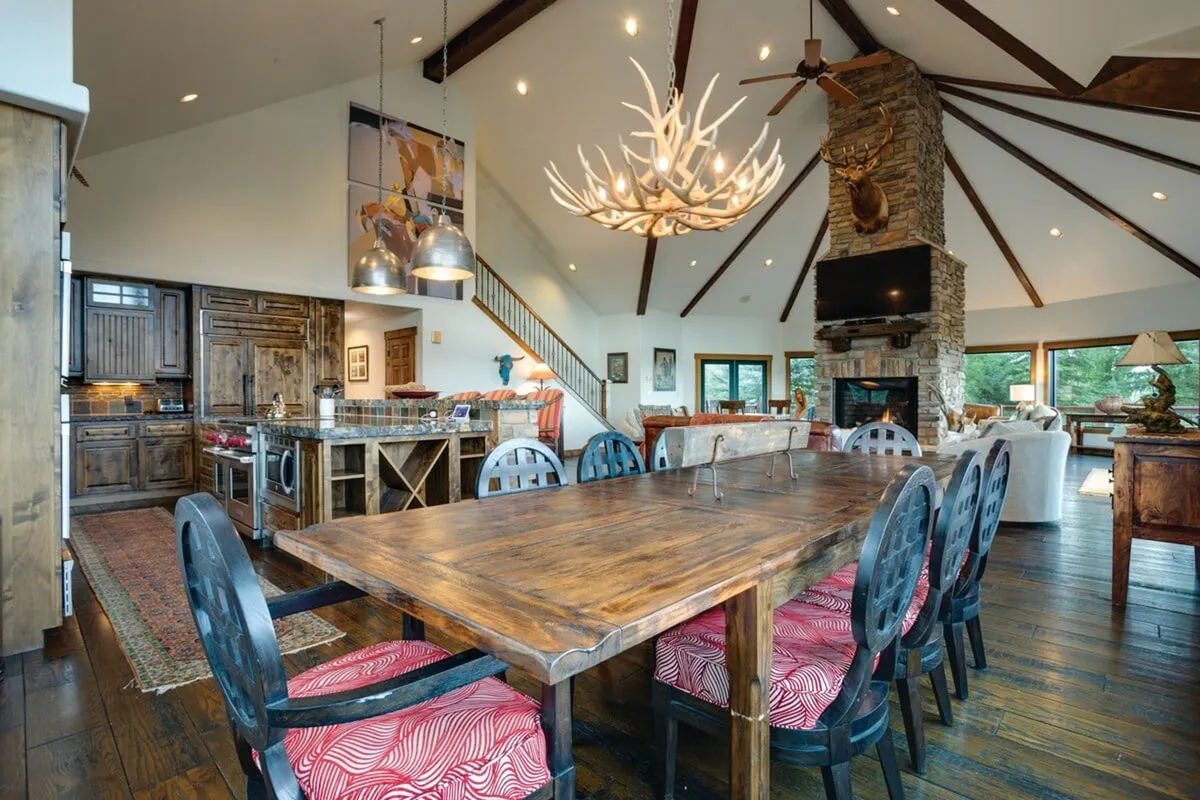 An open-concept space combining a wooden dining table, rustic stone fireplace, antler chandelier, and an open kitchen, all set beneath lofty ceilings. The design creates a seamless transition between eating, relaxing, and sharing time with loved ones.